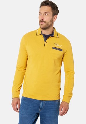 Mann mit kurzen braunen Haaren und Bart trägt ein gelbes Langarm-Poloshirt mit dem Schriftzug „Free Spirit“, blauen Kragenakzenten und blauen Jeans.