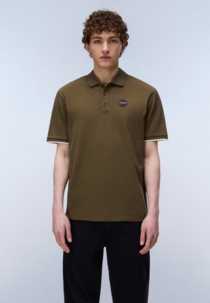 Jeune homme aux cheveux bouclés portant un polo à manches courtes vert olive avec un petit logo rond sur la poitrine et un pantalon noir.