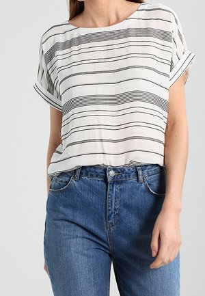 Kobieta w białej bluzce z krótkim rękawem w czarne poziome paski oraz wysokich, niebieskich dżinsach, z rękami po bokach.