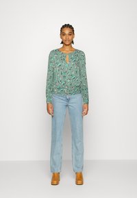 Blusa verde de mangas longas com padrão floral, decote em gota e cintura franzida. Combinada com jeans de perna reta azul claro e tamancos de madeira castanhos.