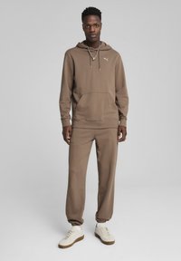 Sudadera con capucha marrón y bolsillo frontal, combinada con joggers a juego. Presenta un tejido suave y puños elásticos, complementados con zapatillas blancas.