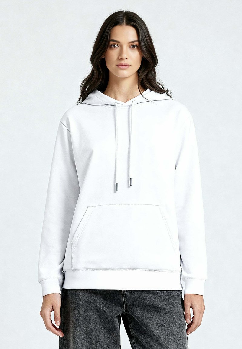 Jeune femme aux longs cheveux noirs portant un sweat à capuche blanc uni et un jean foncé, debout devant un fond gris clair.