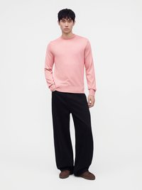 Jeune homme debout portant un pull rose à manches longues, un pantalon noir à jambes larges et des chaussures marron sur un fond blanc uni.