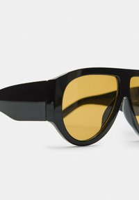 Lunettes de soleil noires avec une monture épaisse, dotées de verres teintés jaunes et d'un design angulaire élégant. Texture lisse avec une finition brillante.