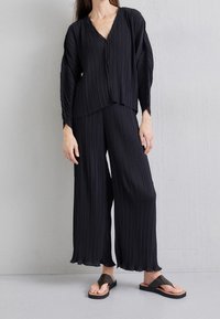 Svart plisserad blus med V-ringning, kombinerad med matchande vida byxor. Skorna har ett mörkt band över en bas av trä.