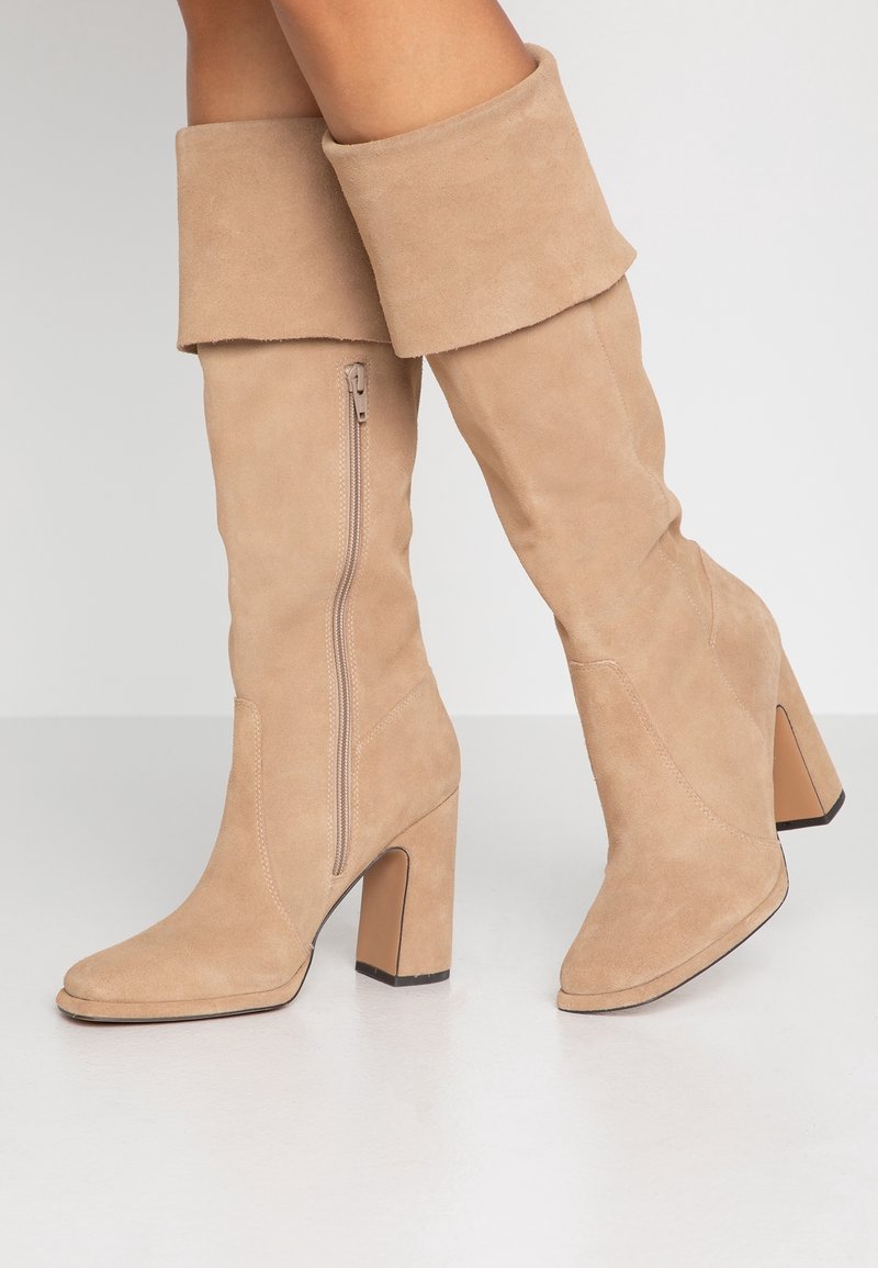 Bottes hauteur genou en daim beige avec un revers, une fermeture éclair latérale et un bout carré, dotées d'un talon bloc. Texture lisse et lignes épurées.