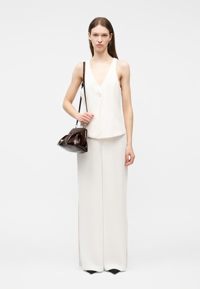 Γυναίκα που φοράει λευκό αμάνικο γιλέκο και παντελόνι με ευρύ πόδι, κρατώντας σκουρόχρωμη καφέ τσάντα ώμου με λουρί, μπροστά σε λευκό φόντο.