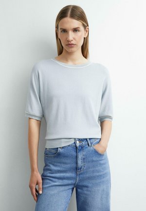 Frau mit glatten blonden Haaren, die einen hellgrauen Kurzarmpullover und blaue hoch taillierte Jeans trägt, steht vor einer weißen Wand.