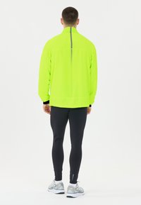 Neonvihreä kevyt takki, jossa on heijastava normaali raita keskellä, yhdistettynä mustiin legginsseihin ja harmaisiin juoksukenkään.