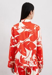 Chemise à motif floral rouge et crème avec des manches longues, un col et des poignets boutonnés. Tissu lisse avec des motifs distincts de feuilles et de fleurs.
