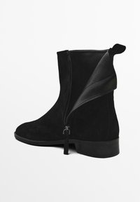 Bottines en suede noir avec un bout arrondi, un petit talon bloc et une fermeture éclair sur le côté. Elles possèdent un accent en cuir flexible près de l'ouverture.