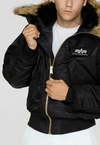 Črna bomberska jakna s kapuco podloženo s sintetičnim krznom, zlatim zadrgo in rebrastimi manšeti. Ima oznako Alpha Industries na prsih.