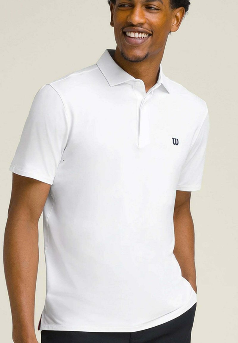 Homme souriant portant un polo blanc à manches courtes avec un petit logo noir en forme de "W" sur la poitrine gauche, debout avec une main dans la poche.