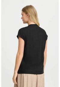 Blusa nera a maniche corte con una superficie liscia e scollatura rotonda, mostrata da dietro, abbinata a una gonna beige fluida.