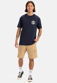 La camiseta de algodón azul marino presenta un diseño de logo redondo en el pecho; combinada con pantalones cortos de carga beige y zapatillas negras con acentos blancos.