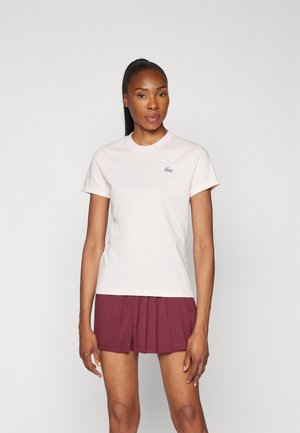 Donna con pelle scura e lunga coda di cavallo nera che indossa una t-shirt rosa chiaro a maniche corte e una gonna plissettata bordeaux, in piedi su uno sfondo neutro.