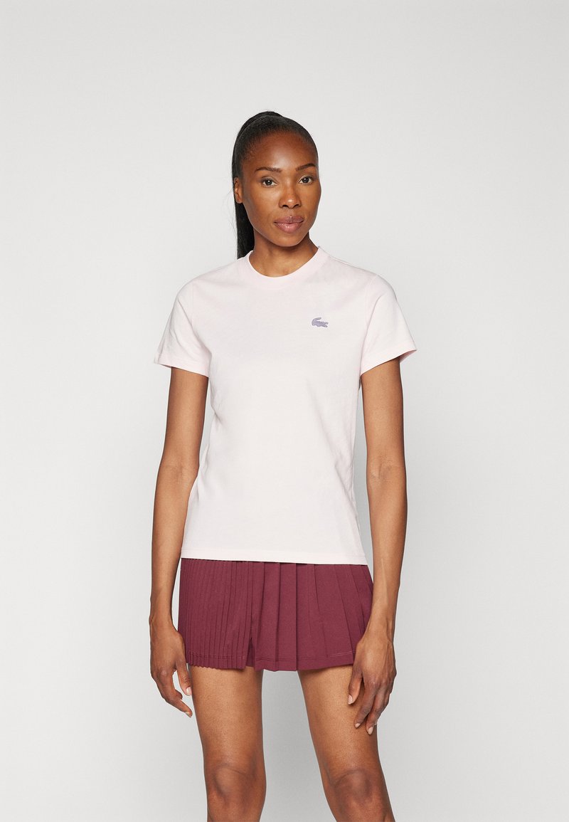 Femme à la peau foncée et longue queue de cheval noire, portant un t-shirt rose clair à manches courtes et une jupe plissée bordeaux, debout devant un fond uni.