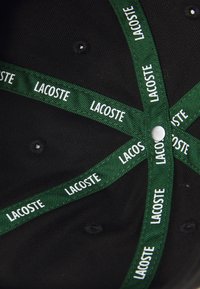 Chapeau noir intérieur avec du ruban en tissu vert portant le logo "LACOSTE" en blanc répété et un bouton central en argent. Surface texturée.