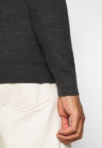 Mörk stickad tröja med ett strukturerat mönster, ribbade ärmar och en åtsittande silhuett, i kombination med ljusfärgade byxor.