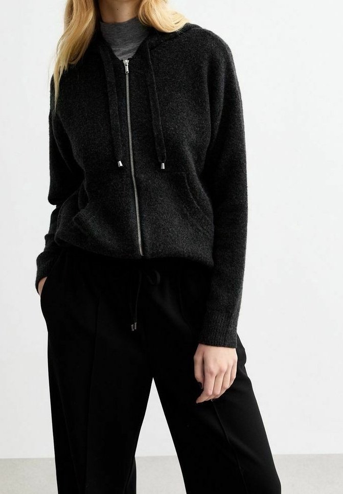 Haut à zip noir en tissu texturé avec cordons de tirage, présentant des poches avant et une coupe décontractée, associé à un pantalon noir.