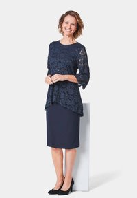 Marineblaues Kleid mit Spitze, drei Viertel langen Ärmeln und asymmetrischem Saum, kombiniert mit einem schlichten Bleistiftrock und schwarzen High Heels.