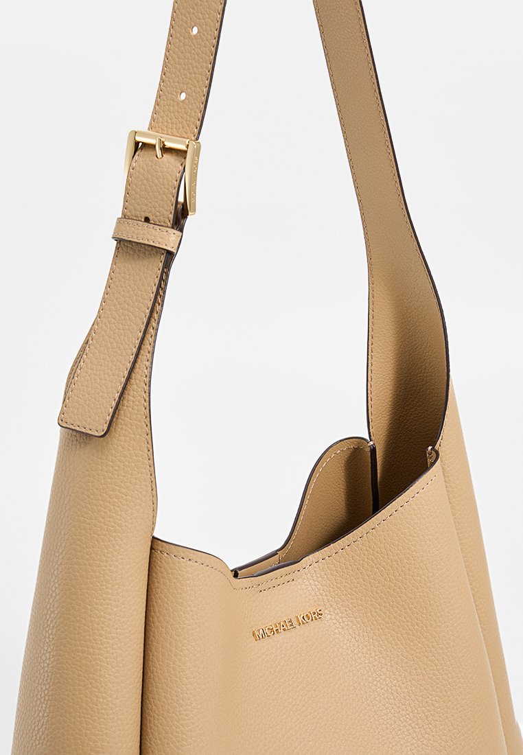 Beige leren schoudertas met een gestructureerde afwerking, voorzien van een goudkleurige gesp en merknaam op de voorkant. Ontwerp zonder deksel met zachte randen.