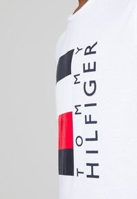 Camiseta blanca de algodón con un logo llamativo en rectángulos azul marino, rojo y negro, con "TOMMY HILFIGER" impreso verticalmente en negro.