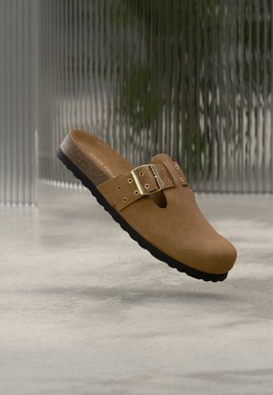 Sabot marron en daim à enfiler avec une bride à boucle ajustable et une semelle noire, présenté flottant au-dessus d'une surface grise avec un arrière-plan flou.