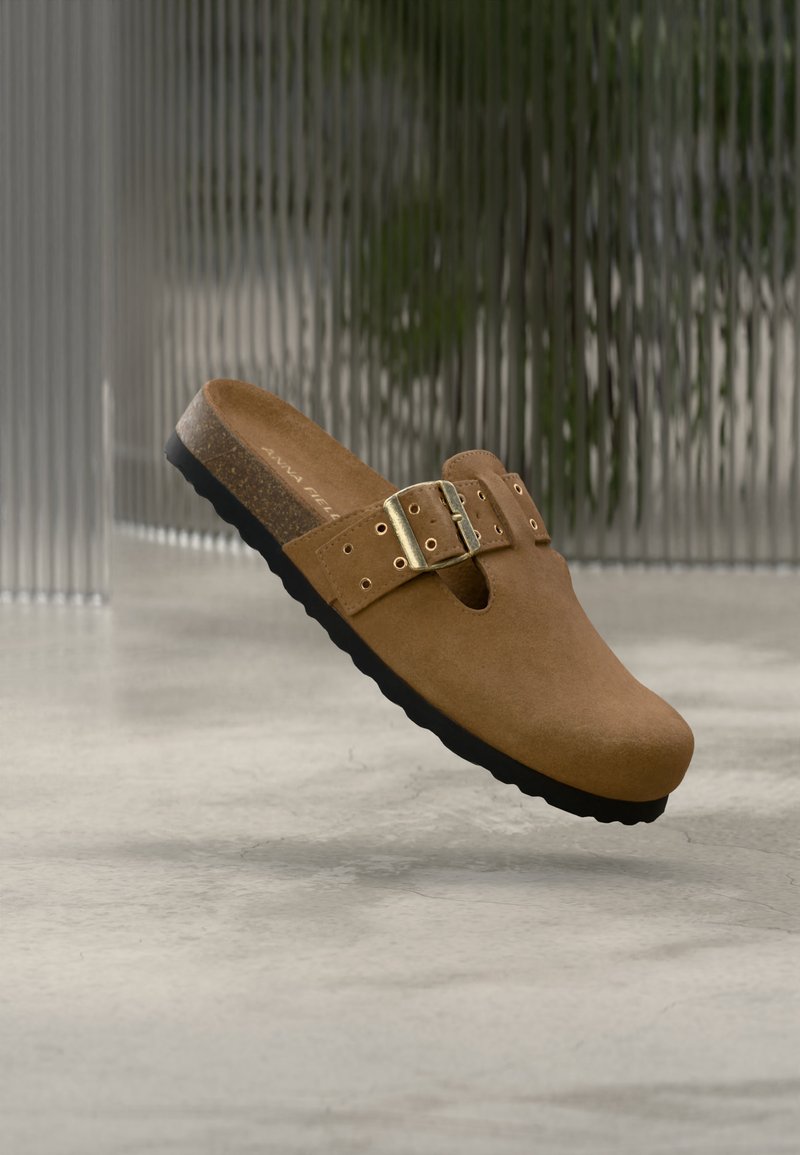 Καφέ κλειστό μοκασίνι σουέτ με ρυθμιζόμενο λουράκι με αγκράφα και μαύρη σόλα, εμφανίζεται να αιωρείται πάνω από μια γκρι επιφάνεια με θολό φόντο.