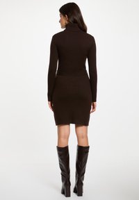 Rippengestricktes, dunkelbraunes Kleid mit Stehkragen, figurbetonter Silhouette und langen Ärmeln, kombiniert mit kniehohen, glänzenden braunen Stiefeln.
