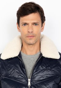 Man met kort bruin haar, gekleed in een marineblauwe jas met een witte fleece kraag en een grijs shirt, die recht vooruit kijkt met een neutrale uitdrukking.