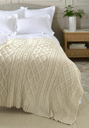 Cremefarbene Kabelstrickdecke, drapiert über ein weißes Bett mit wellenförmigen Kissen neben einem hölzernen Nachttisch mit Büchern und einer Pflanze.