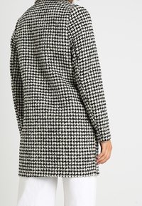 Abrigo de patrón de jaquard en blanco y negro con un ajuste entallado, mangas largas y un cuello, que presenta una tela texturizada y un dobladillo recto.