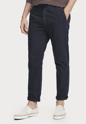 Mężczyzna w podwiniętych granatowych spodniach i białych plecionych butach wsuwanych, stoi z jedną ręką w kieszeni na jednolitym tle.