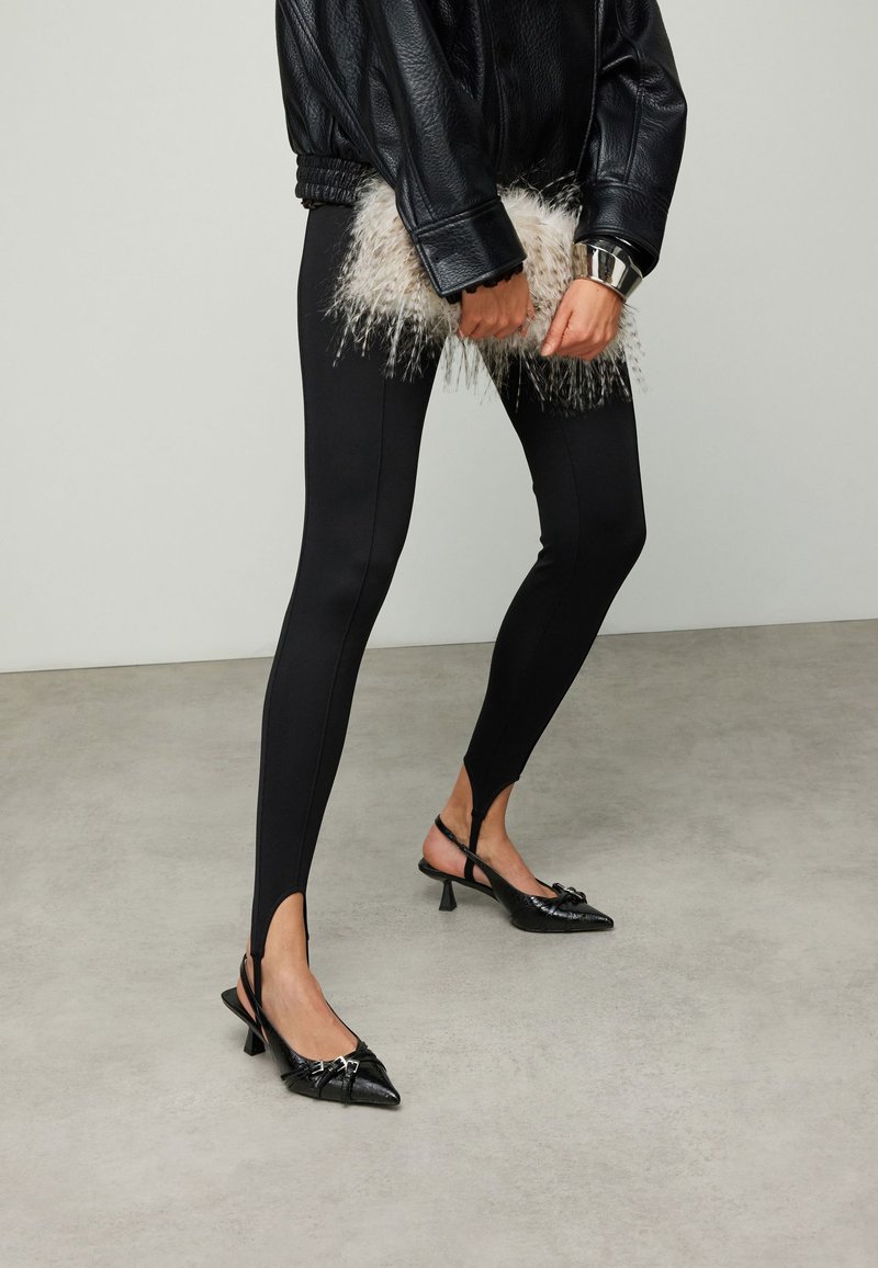 Leggings noirs taille haute avec fentes latérales, assortis à des escarpins noirs à bout pointu et bride arrière, ornés de nœuds décoratifs, et une pochette texturée et duveteuse.