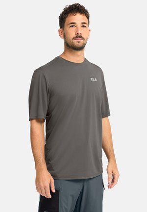 Uomo che indossa una maglietta grigia a maniche corte Jack Wolfskin e pantaloncini cargo blu, in piedi contro uno sfondo chiaro semplice.