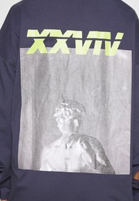 Klassisk staty-byst tryckt på marinblått tyg med stora gul-grön romerska siffror "XXVII" visas ovanför bilden.