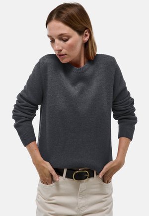 Pull gris foncé à col rond, poignets côtelés et maille texturée. Porté avec un pantalon de couleur claire et une ceinture noire.