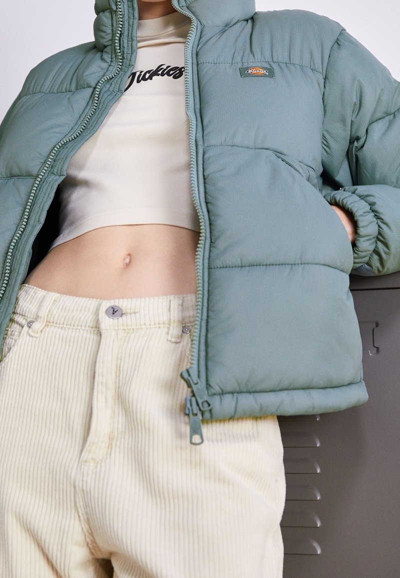 Light green padded jacket with a zipper and hood, worn over a cream crop top and light beige corduroy pants. Textured fabric and slight sheen.