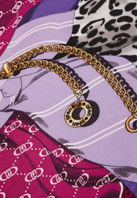 Collana in metallo dorato con pendente rotondo su uno sfondo di tessuto colorato che presenta motivi in viola, rosa e stampa leopardata, con dettagli testurizzati.