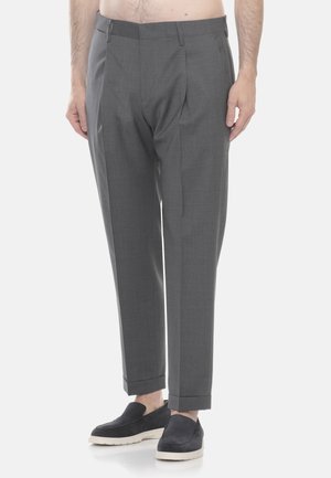 Uomo che indossa pantaloni grigi su misura e scarpe slip-on nere con suola bianca, in piedi davanti a uno sfondo chiaro e uniforme.