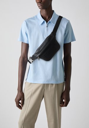 Schwarze Leder-Schultertasche mit geprägt Logo, über der Brust getragen. Hellblaues Kurzarm-Poloshirt mit Reißverschluss, kombiniert mit hellbeigen Hosen.