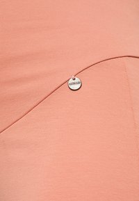 O tecido pêssego com acabamento em relevo apresenta uma costura curva com um pendant prateado marcado com "LOVEZWAT" preso por um fio rosa. Design simples e texturizado.