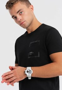 T-shirt in cotone nero con taschino nero texture. Un orologio da polso bianco con quadrante rotondo e dettagli neri a contrasto. 