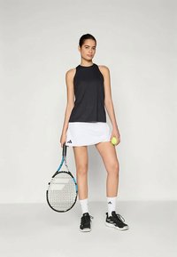 Melnā bez piedurknēm sporta krekls, kas saskaņots ar baltu svārku ar adidas logo. Melnas tenisa apavi, rokās turēts rakete un tenisa bumba.