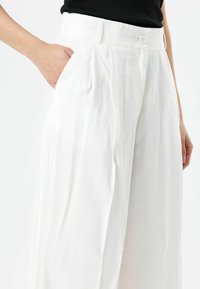 Pantalon blanc à jambes larges avec une taille haute, doté d'une fermeture à boutons et de poches latérales, fabriqué en tissu léger à la texture lisse.