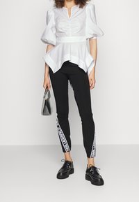 Top bianco con maniche a sbuffo, scollo a V e orlo a peplo, abbinato a leggings neri con stampa del logo laterale e scarpe nere stringate.