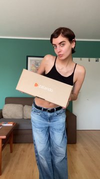 Caixa de cartão castanha com "zalando" impresso em laranja, segurada por uma pessoa vestindo um top crop preto e calças largas de ganga azul claro.