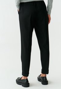 Zwarte, op maat gemaakte broek met een slim fit, voorzien van een omgeslagde zoom en twee achterzakken. Draag met zwarte instapschoenen.