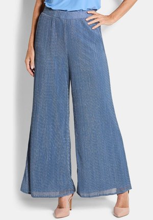 Femme portant un pantalon large bleu à rayures verticales et des escarpins couleur chair à bout pointu, posant avec une main sur la hanche.